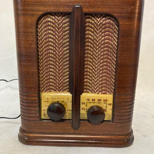 May include: A vertical wooden radio with a polished dark brown finish. It features a textured gold and brown fabric speaker grille with a chevron pattern. Two dark brown circular knobs sit below the grille on a cream-coloured dial panel.