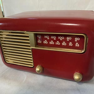 May include: A glossy deep red radio with a gold-toned speaker grille and a matching gold-toned dial frame. The radio features a horizontal frequency display with white numbers ranging from 55 to 160, and two round gold-toned control knobs on the front.