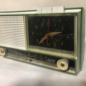 Könnte beinhalten: Ein vintage grünes und weißes Radiowecker mit einem schwarzen Zifferblatt und einem weißen Zifferblatt. Das Zifferblatt hat schwarze Zahlen und einen goldfarbenen Stunden- und Minutenzeiger. Das Radio-Zifferblatt hat schwarze Zahlen und einen weißen Zeiger.