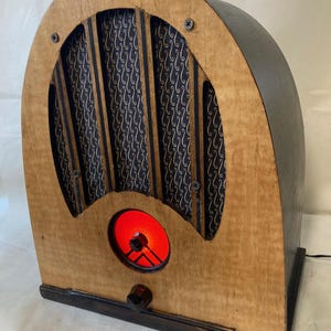 Puede incluir: Una radio de madera de estilo vintage con una rejilla de altavoz con patrón negro y dorado y una esfera iluminada roja. La radio tiene un acabado de madera oscura y una base negra.