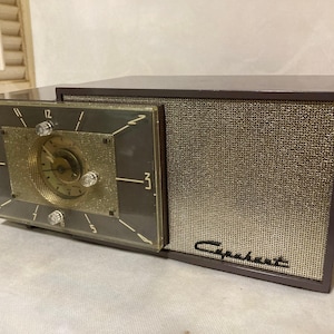 Puede incluir: Una radio Capehart de color marrón oscuro con un reloj analógico integrado en el lado izquierdo. El reloj presenta un centro texturizado dorado, números blancos y perillas transparentes. Una rejilla de malla dorada cubre el lado derecho.