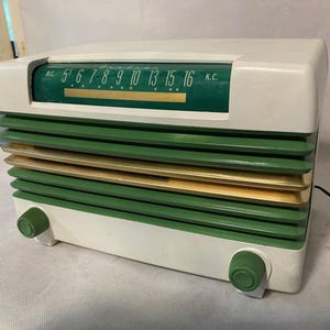 May include: A vintage-style radio featuring a white plastic casing with horizontal green and gold slats. The top section includes a green display panel with white text reading B.C. and K.C. alongside frequency numbers. Two green control knobs are positioned at the bottom left and right corners of the front panel.
