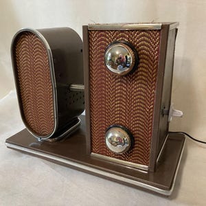 May include: Vintage brown radio with a woven red and gold pattern on the speaker and front panel. The radio has two chrome knobs and a chrome trim. The speaker is oval-shaped and connected to the main body.