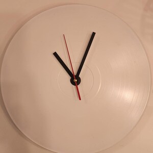 Peut inclure: Une horloge en vinyle blanc avec des aiguilles noires et rouges. Le cadran de l'horloge est blanc avec un centre doré.