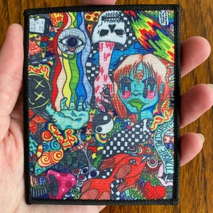 May include: A colorful embroidered patch with a psychedelic design featuring a skull, an eye, a yin yang symbol, a checkered pattern, and a cartoon face with tears. The patch has a black border and is made of fabric.