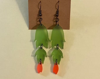 May include: A pair of dangle earrings featuring a layered leaf design. The earrings have a green leaf shape with orange accents and dark metal hooks. The earrings are displayed on a brown card.