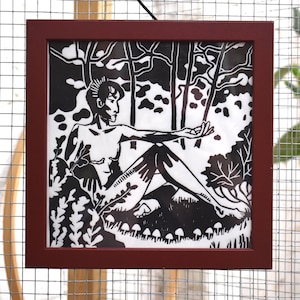 May include: Black and white linocut print in a red frame depicting a person sitting in a forest setting. The person has short, spiky hair and is wearing no clothes. The background features trees and foliage.