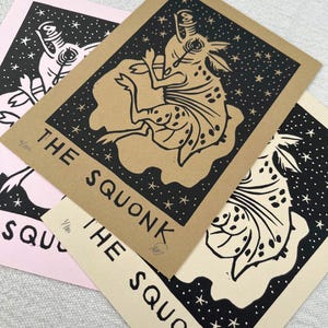 May include: A collection of art prints featuring a whimsical creature design. Each print showcases a black and white illustration of a mythical squonk against a starry background. The prints are on various colored papers, including pink, tan, and cream.