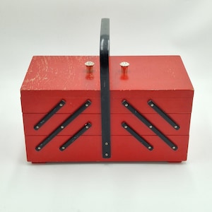 Könnte beinhalten: Eine rote Vintage-Nähkiste aus Holz mit schwarzem Griff und Akzenten. Die Kiste hat eine rechteckige Form mit einem Klappdeckel und mehreren Fächern. Die Kiste hat zwei dekorative Knöpfe auf der Oberseite und schwarze Metallakzente. Die Nähkiste steht auf weißem Hintergrund.