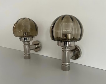 Può includere: Due applique da parete con paralumi in vetro grigio fumé a coste e basi in cromo lucido. Le applique sono montate su una parete bianca e hanno un design classico. Questi apparecchi sono ideali per aggiungere illuminazione ambientale.