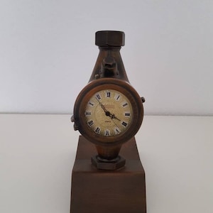 May include: A brown decorative clock with a round face and Roman numerals. The clock is set in a dark brown, rectangular base with a cylindrical top. The clock face has a gold-colored background and black hands.