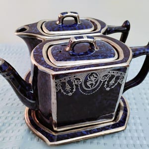 May include: Two matching navy blue teapots with silver trim and handles. The teapots have a square shape with a decorative silver design on the front. The lids have silver handles. The teapots are sitting on a light blue surface.