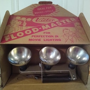May include: A vintage Victor Flood-Master movie lighting kit in its original box. The kit includes three silver-colored floodlights and a black cord. The box is brown with red lettering and features the text "Victor Flood-Master" and "For Perfection in Movie Lighting".
