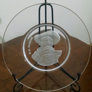 May include: A clear glass plate with a raised, etched image of a man in a hat. The plate is on a black metal stand with a fleur-de-lis design. The text "P.P. RUBENS" is etched on the plate.