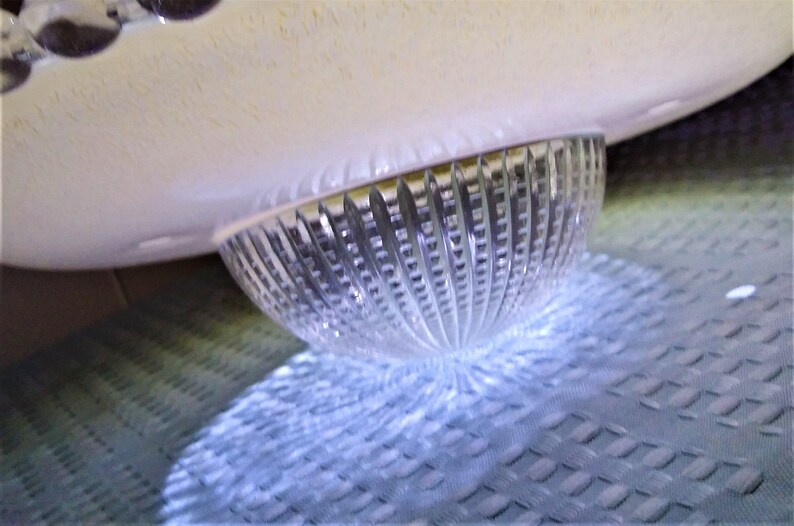 Pu&ograve; includere: Una ciotola di vetro trasparente e scanalato con un bordo arrotondato bianco sporco. La ciotola &egrave; parzialmente illuminata, proiettando un'ombra sulla superficie strutturata sottostante. Il design presenta superfici lisce e strutturate.