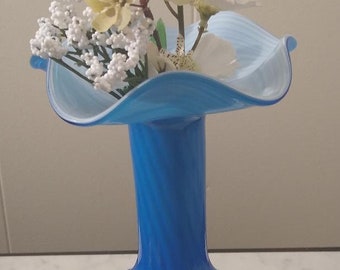 May include: A blue glass vase with a wide, flared opening and a narrow neck. The vase has a white swirl design and is filled with white and yellow flowers.