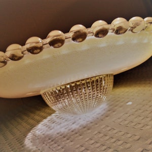 Pu&ograve; includere: Un piatto da portata in vetro vintage con un bordo smerlato di perline rotonde e trasparenti. Il piatto ha una tonalit&agrave; giallo pallido e poggia su una base in vetro trasparente strutturato. Il design ricorda l'arredamento moderno della met&agrave; del secolo.