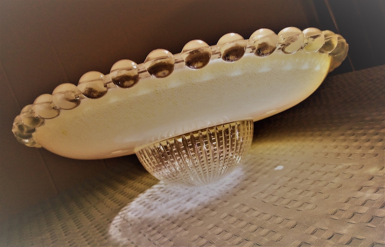 Pu&ograve; includere: Un piatto da portata in vetro vintage con un bordo smerlato di perline rotonde e trasparenti. Il piatto ha una tonalit&agrave; giallo pallido e poggia su una base in vetro trasparente strutturato. Il design ricorda l'arredamento moderno della met&agrave; del secolo.