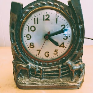 May include: Vintage metal clock with a horseshoe design. The clock face is white with black numbers and hands. The base features two horse figures and a fence. The clock's frame has a bronze finish, and the clock reads approximately 4:10.