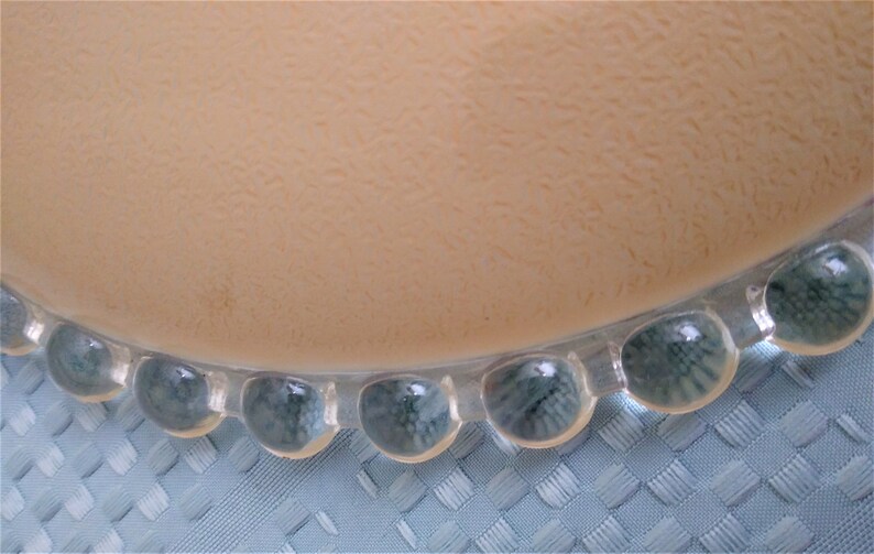 Pu&ograve; includere: Primo piano di un piatto beige con superficie strutturata e bordo trasparente con perline. Le perline hanno una tonalit&agrave; blu-verde. Il piatto poggia su un tessuto blu chiaro intrecciato.