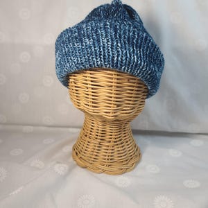 May include: A blue and white knitted beanie hat. The hat is displayed on a wicker head form. The hat has a ribbed texture and a folded brim. The background is a white fabric with a daisy pattern.