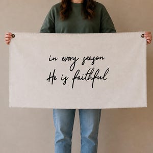 May include: A rectangular off-white fabric wall hanging featuring the black text "in every season He is faithful" printed in a cursive script. The banner includes two dark metal grommets at the top corners for hanging purposes.