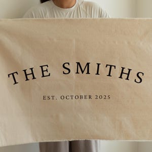 Puede incluir: Alfombra de bienvenida rectangular beige con el texto "THE SMITHS" en negro, en arco, y "EST. OCTOBER 2025" debajo. La alfombra es sostenida por una persona.