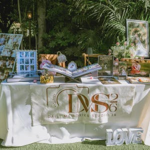 May include: A white table display featuring a large open photo album on a stand, framed wedding photographs, a floral arrangement, and a sign reading Daily Memories Studio 26. White block letters spelling LOVE sit at the front of the table.