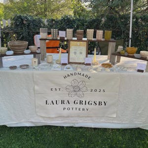May include: A display table covered with a white cloth featuring the text HANDMADE EST. 2025 LAURA GRIGSBY POTTERY and a flower illustration. The table holds various ceramic bowls, mugs, and small pottery items arranged on wooden risers and surfaces.