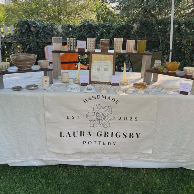 May include: A display table covered with a white cloth featuring the text HANDMADE EST. 2025 LAURA GRIGSBY POTTERY and a flower illustration. The table holds various ceramic bowls, mugs, and small pottery items arranged on wooden risers and surfaces.