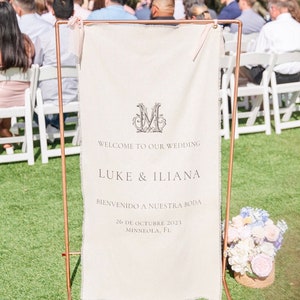 May include: A vertical cream-coloured fabric wedding sign with frayed edges, suspended from a copper-coloured metal frame. The sign features a decorative letter M, the text "WELCOME TO OUR WEDDING", "LUKE & ILIANA", "BIENVENIDO A NUESTRA BODA", and the date.