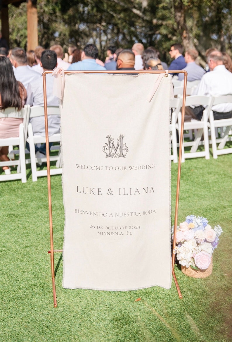 May include: A vertical cream-coloured fabric wedding sign with frayed edges, suspended from a copper-coloured metal frame. The sign features a decorative letter M, the text "WELCOME TO OUR WEDDING", "LUKE & ILIANA", "BIENVENIDO A NUESTRA BODA", and the date.