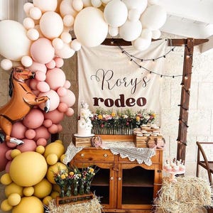Könnte beinhalten: Eine Partydekoration mit einem Ballonbogen in Creme-, Rosa- und Gelbtönen, einem braunen pferdeförmigen Ballon und einem Banner mit der Aufschrift Rory's rodeo. Ein Holzschrank trägt eine Torte, Blumen und Leckereien, ergänzt durch Strohballen.