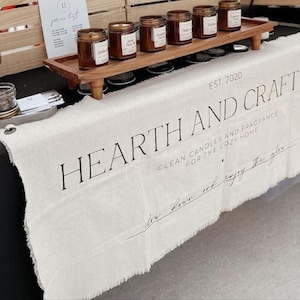 Op de afbeelding: Een crèmekleurige stoffen tafelloper met gerafelde randen voorzien van de zwarte tekst HEARTH AND CRAFT CO. EST. 2020, CLEAN CANDLES AND FRAGRANCE FOR THE COZY HOME, en slow down and enjoy the glow. Een houten verhoger bevat diverse amberkleurige glazen potten.