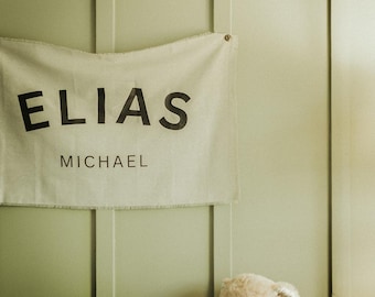Könnte beinhalten: Ein Kinderzimmer mit einem braunen Babybett, einem Teddybär und einem Banner mit der Aufschrift "ELIAS MICHAEL" in schwarzen Buchstaben. Die Wand ist hellgrün gestrichen.