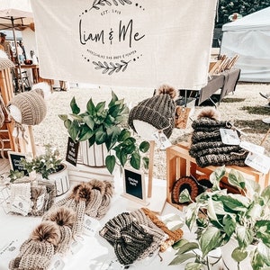 May include: A display of crocheted hats and bonnets with faux fur pom-poms, featuring a banner with the text Liam & Me, Accessories for Baby & Mama, Est. 2018. The items are arranged on a table with green plants in white pots and wooden crates.