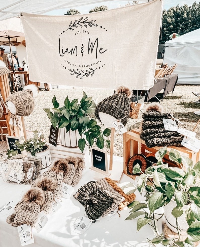 May include: A display of crocheted hats and bonnets with faux fur pom-poms, featuring a banner with the text Liam & Me, Accessories for Baby & Mama, Est. 2018. The items are arranged on a table with green plants in white pots and wooden crates.