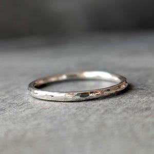 Könnte beinhalten: Ein gehammerter Silberring mit strukturierter Oberfläche. Der Ring ist rund und hat eine leicht unregelmäßige Form, die das Licht reflektiert. Das Schmuckstück ist vor einem grauen Hintergrund platziert.