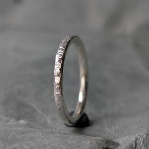 Könnte beinhalten: Ein silberner Ring mit gehammerter Textur. Der Ring ist dünn und hat eine leicht unregelmäßige Oberfläche, die das Licht reflektiert. Der Ring ist vor einem grauen Hintergrund platziert, der seinen metallischen Glanz hervorhebt. Ein Schmuckstück.