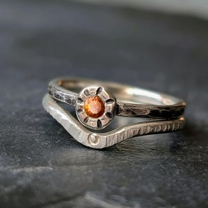 Könnte beinhalten: Zwei Silberringe. Der obere Ring hat ein strukturiertes Band und einen zentralen orangefarbenen Edelstein. Der untere Ring hat eine Chevron-Form mit einer strukturierten Oberfläche. Die Ringe sind übereinander gestapelt.