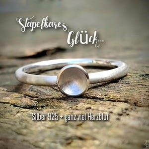 Könnte beinhalten: Ein Silberring mit einem runden, erhabenen Mittelstein. Der Ring ist vor einem rustikalen Holzuntergrund platziert. Der Text auf dem Bild lautet "Stapelbares Glück..." und "Silber 925 + ganz viel Herzblut".