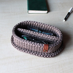 May include: A handmade, oval-shaped, crochet basket in shades of brown, holding two pairs of eyeglasses. A small leather tag is attached to the side. A pen and a green book are in the background.