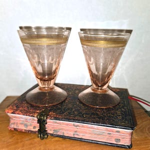 Puede incluir: Dos copas de cristal rosa con un borde ancho y acampanado y una base circular resistente. Cada copa presenta una banda dorada horizontal cerca de la parte superior y un intrincado patrón floral grabado que cubre la parte superior del cuenco.