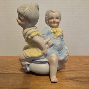 Könnte beinhalten: Eine Porzellanfigur, die zwei Kinder darstellt. Eines trägt ein gelbes Kleid, das andere einen blauen Anzug. Die Kinder sitzen auf einem weißen Sockel mit blauen Akzenten. Die Figur hat eine Vintage-Ästhetik.