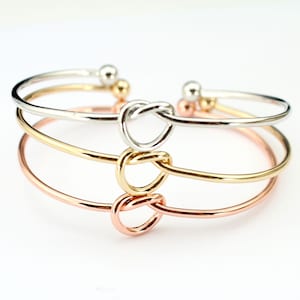 May include: Three delicate bangle bracelets with a knot design. The bracelets are silver, gold, and rose gold.