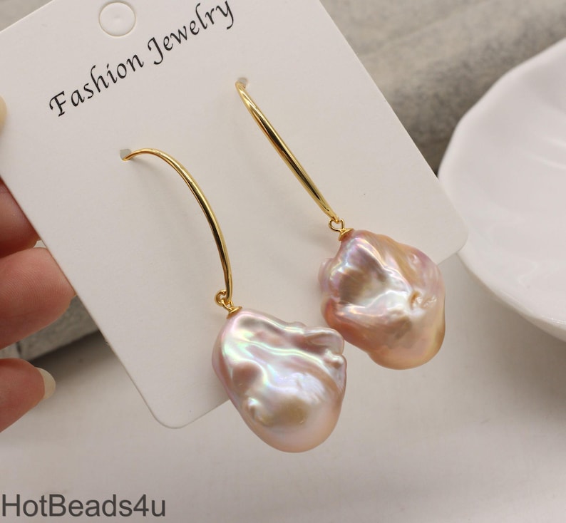 May include: A pair of gold hoop earrings with a large, irregular pink pearl dangling from each hoop. The pearls have an iridescent sheen.