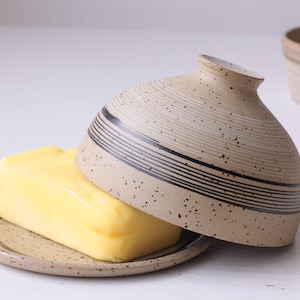 Op de afbeelding: Een botervlootje van keramiek met een deksel en een basis. Het deksel heeft een bruin en zwart gestreept ontwerp. De basis heeft een vergelijkbaar ontwerp. Een klompje boter ligt op de basis.