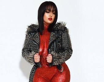 May include: A woman in a red sequined jumpsuit and black studded jacket. The jacket has silver spikes and a belt. She is wearing black heels with gold studs. The background is white.