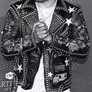 Pode incluir: Uma jaqueta de couro preta com tachas prateadas e detalhes em forma de estrela. A jaqueta tem bordados florais nas mangas e uma camiseta branca por baixo. A pessoa está usando calças com padrão preto e branco.