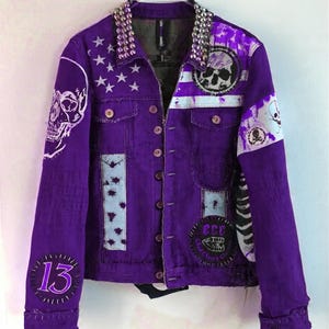 May include: A purple denim jacket with silver stud detailing on the collar. It features white skull graphics, patches, and the number "13". The jacket has a button-up front and a distressed punk rock style.